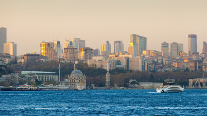 Panoramic view of Istanbul, Turkey, showing modern towers and traditional architecture along the Bosphorus during sunset. Ferries and boats are visible on the water in the foreground.