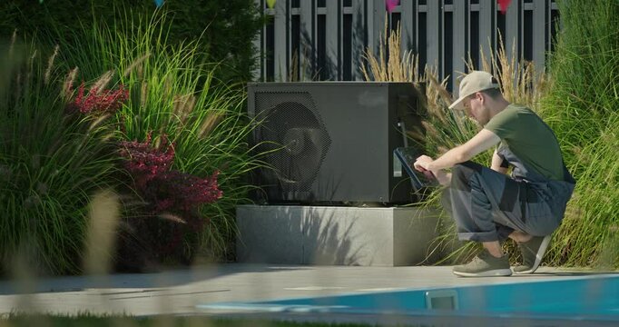 Young technician squatting near poolside heat pump with tablet, surrounded by plants and colorful flags, performing outdoor maintenance work