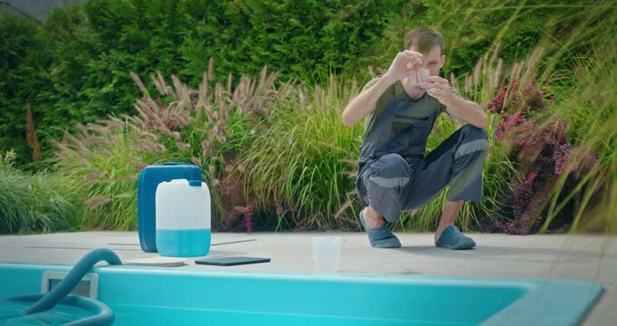 Pool technician squats beside a swimming pool while examining water quality using a color test kit, with chemical containers and maintenance tools nearby.