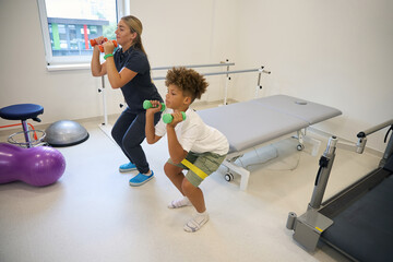 Teenage boy and young physiotherapist, perform squat exercises together © Svitlana