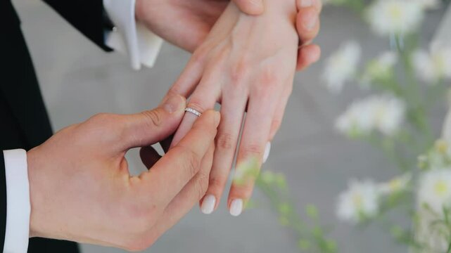 The groom puts a ring on his bride's hand. The camera takes a close-up of the newlyweds' hands