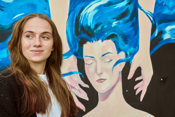 Young adult female with brown hair stands beside colorful canvas. Mural shows stylized face with...