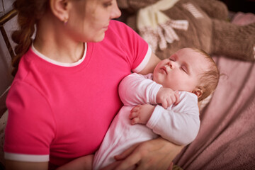 Young woman with dark hair in pink shirt holds sleeping baby girl. Infant in white clothing rests peacefully, enveloped by warm indoor lighting. Soft background create cozy atmosphere