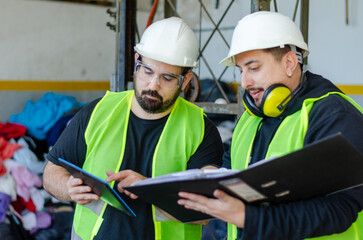 Two male industry factory workers wearing safe gear and holding a binder and a digital tablet....