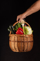 Male hand holding a wicker basket full of healthy colorful vegetables on a black background