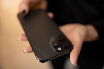 Close-up of unrecognizable woman holding broken smartphone with cracked back glass, checking damaged surface with finger. Frustrated female touching cracked damaged back glass of phone.