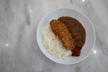 Japanese katsu curry with crispy breaded pork cutlet, white rice, savory curry sauce with vegetables, and pickled ginger, presented in a white bowl on a marble surface.