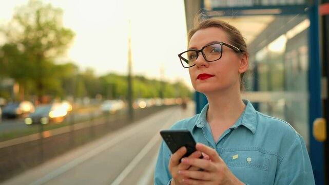 Woman stands at a transport stop, using smartphone and waiting for the tram.