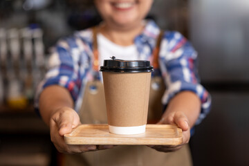 Smiling Asian barista in apron holding wooden tray with takeaway coffee cup toward camera. Focus on hand and cup, background blurred. Warm customer service concept in cozy café environment.