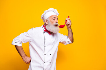 Senior chef in uniform tasting food with red spoon on vibrant yellow background, concept of...