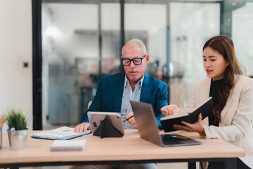 Senior manager and young assistant discussing a project while collaborating with a laptop, tablet, and notebook in a bright, modern office setting. Engaging in teamwork and strategic planning