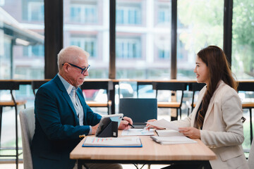 Senior businessman and young businesswoman discussing and working together, using tablet, laptop and paperwork, sitting at desk in modern office space with large windows