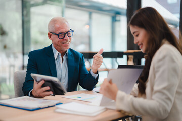 Obraz premium Senior manager showing thumbs up to young businesswoman during a meeting, expressing his appreciation for her work, while reviewing documents and data on a tablet and laptop in the office