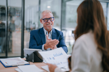 Senior business manager engaging in a discussion about marketing results with a female colleague during a meeting in a modern office, utilizing charts and a tablet for analysis