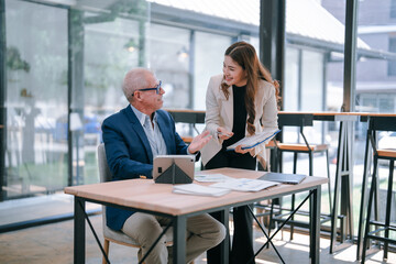 Senior manager sitting at his desk discussing with his young assistant about business strategy using a digital tablet and some documents in a luminous office