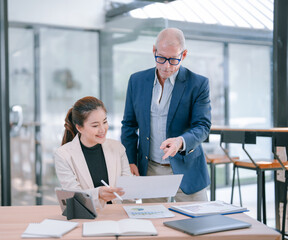 Senior manager coaching a young Asian businesswoman while reviewing paperwork, pointing at a...