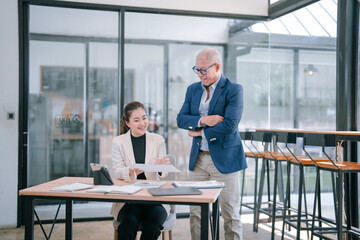 Asian businesswoman presenting a document to a senior businessman while discussing marketing strategies during a meeting in a modern office environment filled with natural light