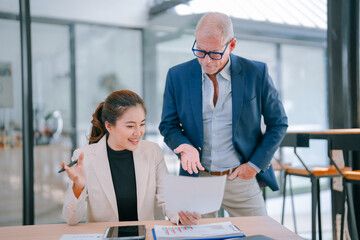 Senior manager coaching young Asian businesswoman in modern office, reviewing paperwork and discussing financial reports to enhance business strategies and teamwork