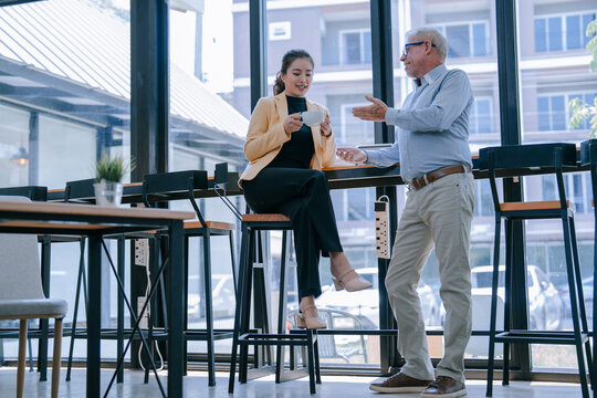 Asian businesswoman sitting and enjoying coffee while engaging in a discussion with a senior businessman during a coffee break in a modern office cafeteria, fostering collaboration and teamwork - Powered by Adobe