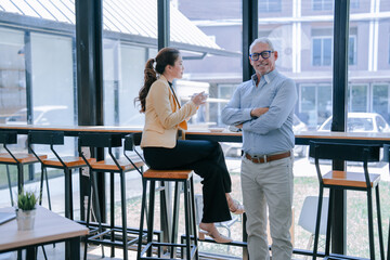 Senior manager with crossed arms and businesswoman drinking coffee are having a conversation during a break in a modern office space with large windows