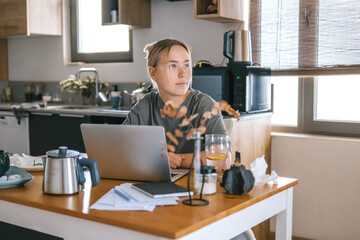  Gen Z woman working from home at cozy kitchen table with laptop and phone. Digital nomad...