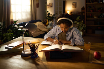 Young woman sitting at desk with open book, wearing headphones, and concentrating on studying late...