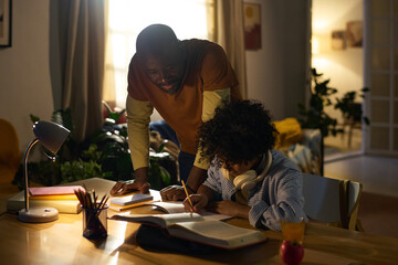 African American man standing next to child while assisting with homework, both engrossed in study in cozy home environment lit with warm lights, showcasing family bond