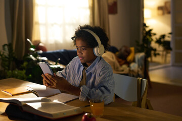 Young African American woman concentrating on her studies at table with tablet and headphones. Cozy home environment with open books and warm ambient lighting creating focused atmosphere