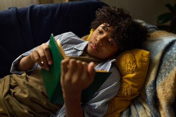 Young boy deeply engrossed in book while lying on couch with pillows surrounding him portraying...
