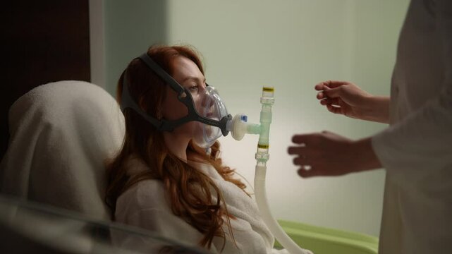 Doctor assisting putting on oxygen mask for respiratory training to female patient in bathrobe during medical procedure in hospital or clinic room. Procedure revitalizing and anti-aging effect.