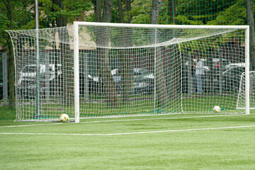 A soccer goalpost stands on a vibrant green field, with two yellow and white balls resting nearby. Trees provide shade as players prepare for a game on a sunny afternoon