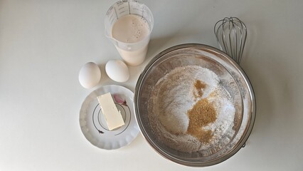 Flat lay of baking ingredients including flour, sugar, butter, eggs, milk, and a whisk on a white surface. Concept of home cooking, ingredient simplicity, and clean food preparation