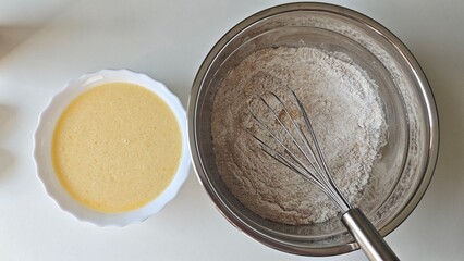 Bowl of flour and whisk next to a bowl of liquid waffle batter on a white surface. Concept of homemade cooking, preparation process, and baking simplicity