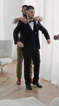 Vertical shot of two biracial best men helping groom with black suit, adjusting bowtie and bouto