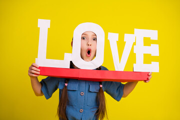 Adorable girl holding a love sign on a vibrant yellow background, expressing surprise and joy while dressed in a trendy outfit.