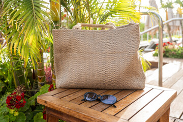 Stylish woven beach bag and black sunglasses rest on a wooden table surrounded by tropical plants at a sunny resort, evoking a relaxed vacation atmosphere.