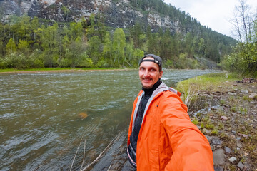 An Adventurous Man Dressed in a Bright Orange Rain Jacket Stands Confidently by the Tranquil Riverbank, Ready to Embark on His Next Journey into the Wilderness and Explore Natures Beauty