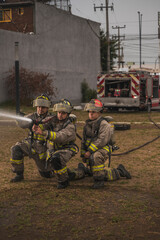 Fototapeta premium Three brave firefighters, standing behind a curtain of water, extinguishing a fire, with the tanker truck behind them.