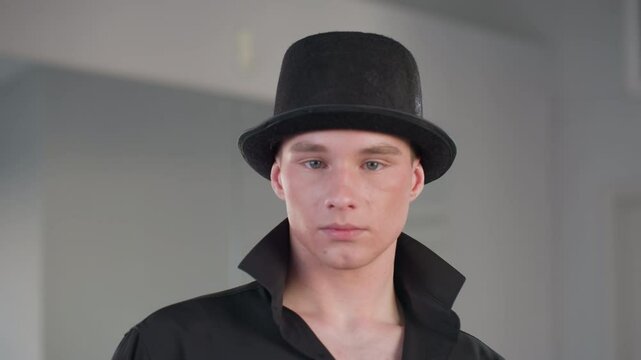 young man in black outfit and top hat stares confidently into camera inside loft studio with mirror reflections highlighting neck tattoo and popped collar exuding modern style and suave attitude