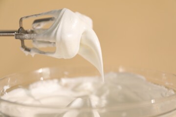 Hand mixer with whisks and bowl of whipped cream on beige background, closeup