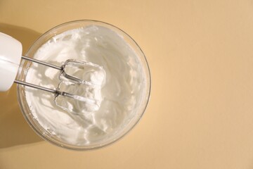 Hand mixer with whisks and bowl of whipped cream on beige background, top view. Space for text