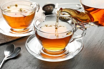 Pouring herbal tea from teapot into cup at black table, closeup