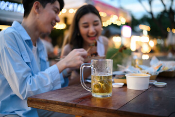 A beer glass sits in focus as two friends laugh and enjoy street food together, capturing a lively and relaxed evening at an outdoor night market