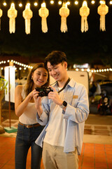 Two friends happily reviewing photos on a camera under decorative lantern lights at a night market, enjoying their travel experience together