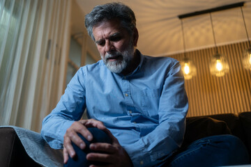 Fototapeta premium Old bearded man suffering from strong knee pain while sitting in living room during evening. Senior male feeling sharp joint ache, arthritis or injury discomfort.