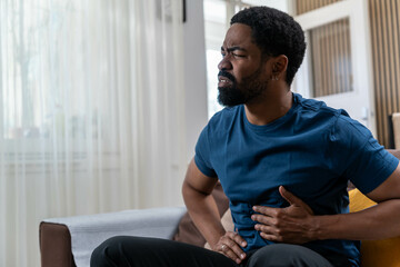 Sad young Black man suffering from strong stomach ache while sitting on couch at home during...