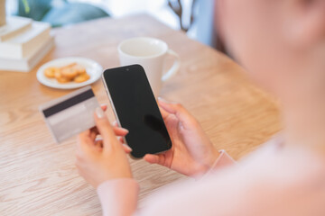 A beautiful young Asian woman smiles as she enjoys a cup of coffee in her cozy kitchen, using a smartphone and browsing a mobile app while relaxing at home