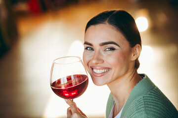 Charming young woman enjoys a glass of red wine in a cozy indoor setting, radiating a happy and relaxed atmosphere