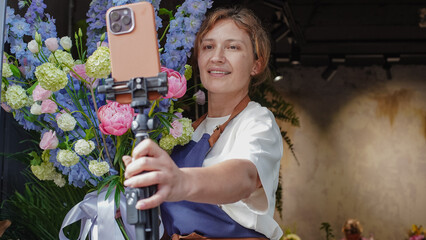 Florist takes a selfie on her phone with a bouquet she has assembled. Florist conducts online...