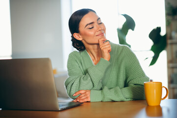 Young brunette woman enjoying leisure time at home in a cozy sweater, sitting at a desk and smiling peacefully indoors